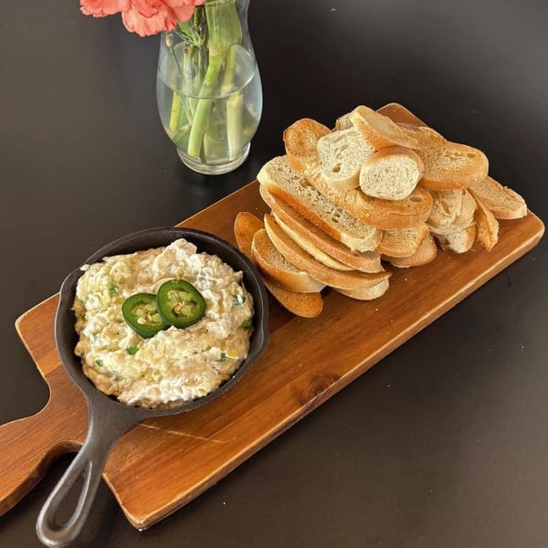 Bruchelle's Bagel Bistro's creamy dip with jalapeños in a skillet, served with sliced baguette on a wooden board.